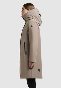 Beige vandtæt frakke med høj krave og justerbar hætte. Har lynlåslommer på siden og et lille logo-patch i kanten.