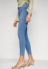 Jeans skinny de mezclilla azul con cintura de altura media, largo recortado y desgastes sutiles, combinados con tacones altos beige con tiras.