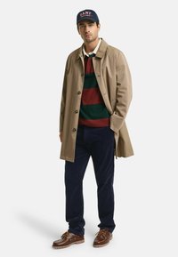 Beige Trenchcoat über einem rot-grün gestreiften Pullover, marineblaue Cordhosen, braune Lederschuhe und einer marineblauen Mütze mit roter Schrift.