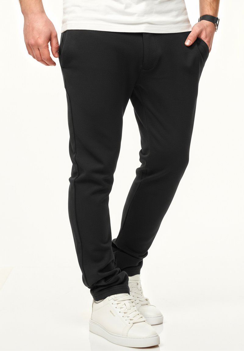 Schwarze Jogginghose aus weichem Stoff, mit Seiten Taschen, schmal zulaufenden Beinen und elastischen Bündchen. Kombiniert mit weißen Turnschuhen.