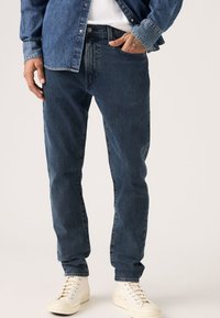 Tamsiai mėlyni džinsai iš džinsinio audinio, laisvo silueto, su penkių kišenių dizainu ir subtiliu nusidėvėjimo efektu. Dėvimi kartu su baltu marškinėliu trumpomis rankovėmis ir sportbačiais.