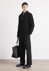 Suéter negro de canalé con collar con cremallera, combinado con pantalones cargo negros con bolsillos laterales y un bolso tote negro. Zapatos de cuero lisos.