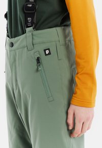 Groene broek van gladde stof, voorzien van verstelbare banden, een ritszak en een logo patch. Complementair met een oranje shirt met lange mouwen.