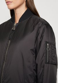 Černá bomber bunda s předním zipem, žebrovaným límcem a bočními kapsami se zipem na levém rukávu. Hladká, lesklá tkanina.