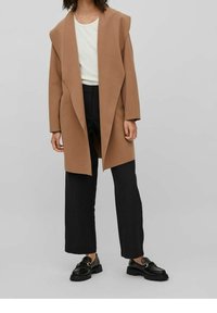 Manteau marron surdimensionné au design drapé, porté sur un haut blanc et un pantalon large noir. Des mocassins noirs complètent la tenue.