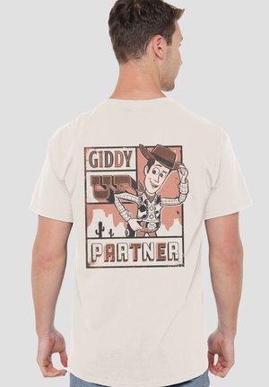 Man wearing beige t-shirt with cartoon cowboy tipping hat and text "Giddy Up Partner" on the back against plain background.