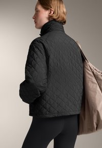 Giacca trapuntata nera con colletto alto, motivo testurizzato. Una borsa beige chiaro è appoggiata sulla spalla. Lo sfondo è neutro e discreto.