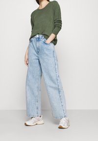 Grüner Strickpullover, hoch taillierte, hellblaue Jeans mit weitem Bein und weiße Sneaker mit roten Akzenten. Glatte Textur und entspannte Passform.