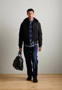 Black bomber jacket over a denim shirt, blue jeans, and black shoes. Model holds a dark leather bag. Smooth texture, casual design.