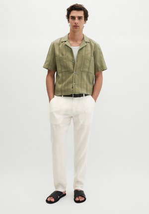 Junger Mann steht mit den Händen in den Taschen weißer Hose, trägt ein olivgrünes Kurzarmhemd, schwarzen Gürtel und schwarze Sandalen vor hellem grauem Hintergrund.