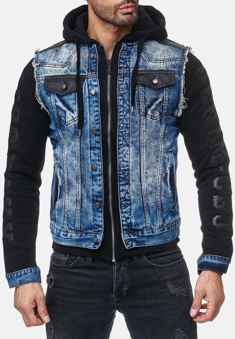 Blauwe spijkerjas met zwarte sweatshirtmouwen en capuchon. Heeft een verweerd randje, drukknoopsluiting, borstzakken en de tekst "Proudly" op de mouw.