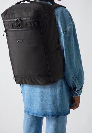 Person trägt einen großen schwarzen Rucksack mit mehreren Reißverschlussfächern über einer blauen Jeansjacke und Jeans.