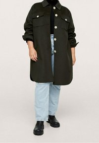 Chaqueta de camisa oversized de color verde oscuro con dos bolsillos en el pecho, grandes botones y mangas enrolladas, combinada con unos jeans azul claro con puños y botas negras.