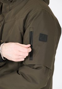 Olivgrön isolerad jacka med en dragkedjeförsedd ärm ficka och en svart logotyp patch. Materialet verkar slätt med en något glänsande yta.