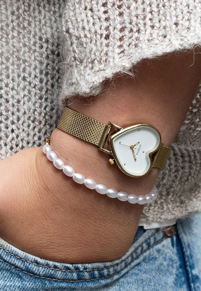 Montre avec bracelet en maille dorée et cadran blanc en forme de cœur, ornée d'accents dorés, associée à un bracelet en perles de nacre blanche sur un poignet contre un tissu tricoté.