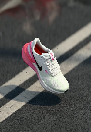 Chaussure de course Nike blanche et rose avec un swoosh noir, flottant au-dessus d'une route asphaltée avec des marquages blancs de voie.