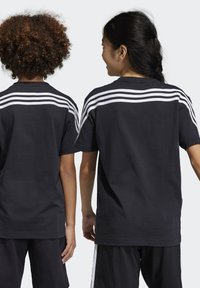 Camisetas de algodón negro con diseño de tres rayas blancas a lo largo de los hombros, exhibidas en dos figuras desde la parte de atrás.