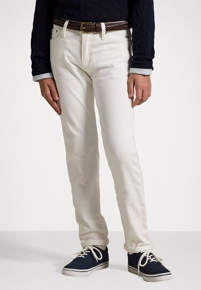 White cotton pants with a straight cut, featuring a braided brown leather belt and worn with navy sneakers. Textured fabric with a clean design.