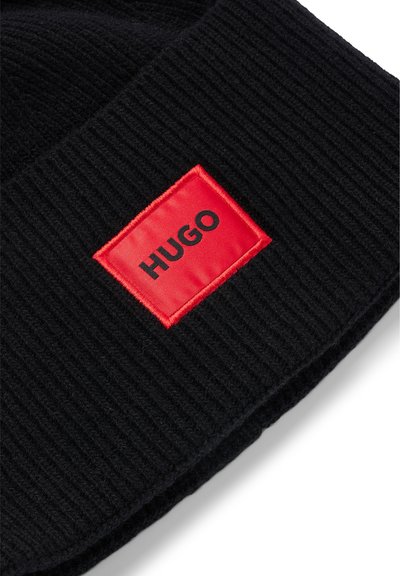 Beanie noir à côtes avec un badge rectangulaire rouge arborant le mot "HUGO" en noir. Texture douce, ajustement serré, adapté aux temps froids.