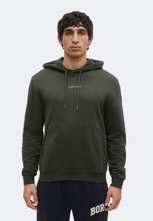 Man draagt een donkergroene hoodie met voorzak en tekst op de borst, gecombineerd met marineblauwe broek met witte tekst op de linker dij.