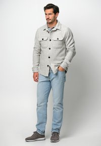 Grau strukturiertes Hemdjackett mit Kragen, zwei Brusttaschen und schwarzen Knöpfen, kombiniert mit hellblauen Jeans und grauen Sneakern.