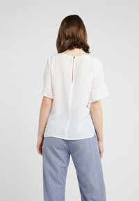 Weißes, kurzärmeliges Blusenshirt mit rüschierten Kanten und einem kleinen Knopfverschluss hinten, aus leichtem Stoff. Kombiniert mit karierter Hose.