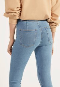 Högmidjade blå denimjeans med två bakfickor, sömnadsdetaljer och en figursydd design. Tyget har en lätt textur som är typisk för denim.