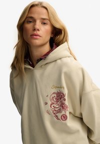Blonde vrouw die een beige hoodie met rode draakborduursel draagt en een rood geruiten overhemd eronder, kijkt recht vooruit met een neutrale uitdrukking.