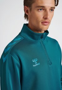 Camiseta deportiva de media cremallera en color verde azulado, hecha de tejido suave; cuenta con un alto cuello, acentos en forma de flecha azules en los hombros y un logo en el pecho.