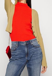 Col roulé rouge côtelé sous un cardigan boutonné beige clair, associé à un jean en denim bleu et un petit sac bandoulière rouge.