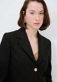 Czarna marynarka o strukturalnym fasonie, z jednym złotym guzikiem i klapami; noszona na prążkowanej beżowej bluzce. Gładka tekstura materiału.