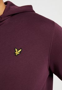 Mörkröd hoodie med en liten broderad fågelikon i gult på vänster bröst, som visar tygstruktur och sömnadsdetaljer.