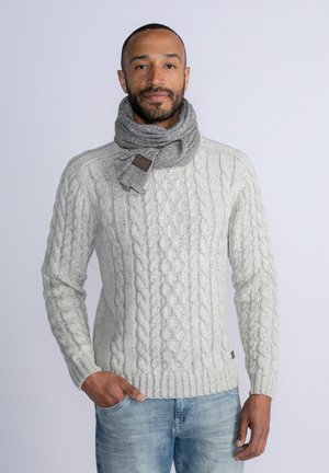 Suéter de punto gris claro con un patrón de trenzas, combinado con una bufanda tejida de un gris más oscuro que cuenta con una etiqueta de cuero. Modelo vistiendo jeans azules.