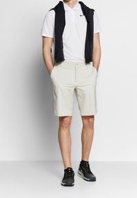 Pánská bílá polo tričko, světle béžové šortky, černá bezrukávová bunda přehozená přes ramena a černé sportovní boty s zelenými detaily.