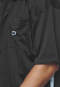 Zwart shirt met korte mouwen en een borstzakje. Heeft een blauw "P"-logo op het zakje en lichtgekleurde accenten aan de zijkanten. Gladde textuur.