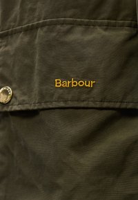 Κοντινή  λήψη σκούρου πράσινου υφάσματος με το λογότυπο Barbour κεντημένο με κίτρινη κλωστή και μια μεταλλική κουμπί πατιλέτα στη αριστερή πλευρά.
