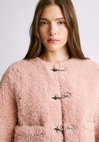 Rosa, flauschiger Schaffellmantel mit rundem Kragen, zwei vorderen Taschen und Metallverschluss zum Schließen. Weicher Stoff und gemütliches Design.