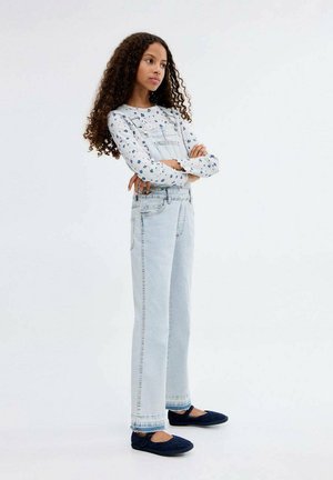 Hellblaue Denim-Trägerhosen mit geradem Schnitt, ausgestattet mit einem verwaschenen Look, Taschen und einem weißen Langarmoberteil mit blauen Blumenmustern. Marineblaue Ballerinas.