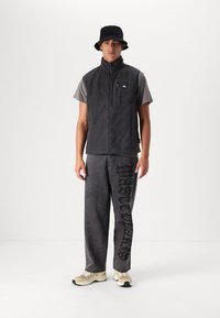 Gray fleece vest with a zipper and front pocket, paired with gray pants featuring black lettering, and beige sneakers; worn with a black bucket hat.