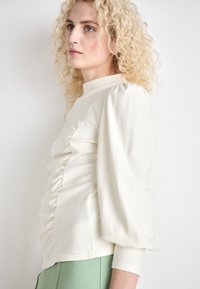 Cream-colored, long-sleeve top with ruched sides and puffed sleeves, featuring a high collar. Paired with green trousers.