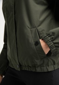 Oliwkowa kurtka z zapięciem na zamek, elastycznymi mankietami i boczną kieszenią z detalem w postaci guzika. Gładki materiał o delikatnym połysku.