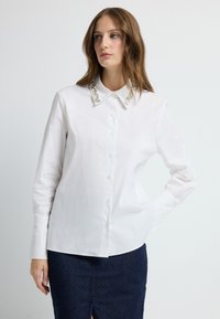 Witte blouse met een decoratieve kraag met heldere versieringen, gecombineerd met een donkerblauwe textuurrok. Lange mouwen, klassieke pasvorm.