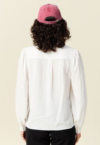 Weißes Langarmhemd mit weichem Stoff, ausgestattet mit Puffärmeln und einem yoke-Design auf der Rückseite. Getragen mit einer verblassten rosa Kappe.