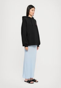 Mulher em pé com um hoodie oversized preto, uma saia até os tornozelos de cor azul clara e sandálias pretas contra um fundo branco liso.