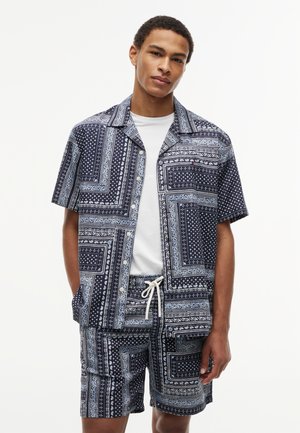 Young man wears patterned black and white short-sleeve button-up shirt and matching shorts with white drawstring over white t-shirt.