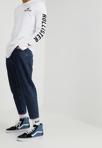 Långärmad vit skjorta med svart text, blå denim smalrandiga jeans och svart/blå hög-top sneakers med vita snören och randiga strumpor.