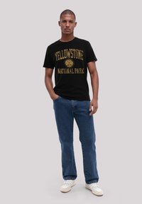Black t-shirt featuring gold print of "Yellowstone National Park," paired with blue denim jeans and white sneakers. Simple and casual design.