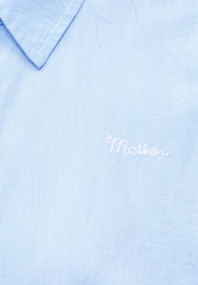 Ανοιχτό μπλε πουκάμισο με γιακά, με την λέξη «Mother» κεντημένη με λευκή κλωστή στο αριστερό τμήμα του στήθους.