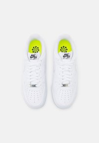 Ett par vita Nike Air Force 1-sneakers med vita snören och neongula innersulor med en svart blomliknande logotyp, sett ovanifrån.