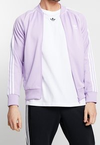 Ljuslila sportjacka med vita ränder, dragkedja och ribbade manschetter, bärs över en vit t-shirt och svarta byxor.